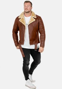 Giacca di pelle marrone con colletto in pelliccia sintetica, maniche trapuntate, tasche con zip e vita con cintura. Indossata sopra una T-shirt bianca e jeans neri.