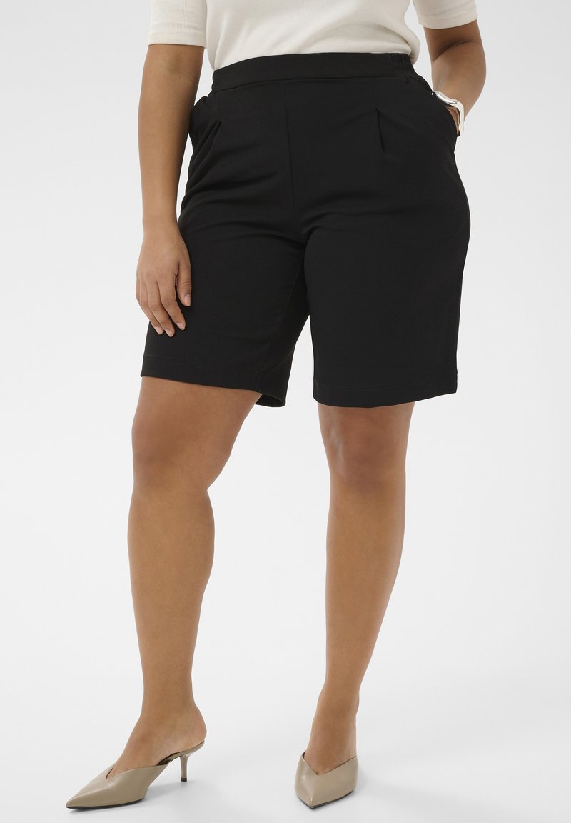 Femeie purtând pantaloni scurți negri, croiți, bej cu vârf ascuțit și un top alb, cu o mână în buzunar, pe un fundal simplu.