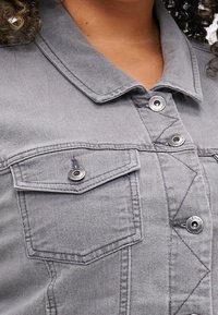 Šedá džínsová bunda s klasickým límcem, dvěma kapsami na hrudi, stříbrnými knoflíky a viditelnými švy. Texturovaná látka.