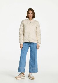 Licht beige gewatteerde jas met een ronde kraag, drukknoopjes en zijzakken. Gecombineerd met wijde blauwe spijkerbroeken en sportieve sneakers.