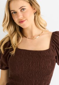 Braune, geraffte Bluse mit kurzen Puffärmeln und quadratischem Ausschnitt. Der strukturierte Stoff ist weich und hat dezente Details. Einfaches goldenes Halskettchen.