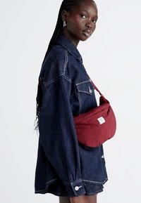 Borsa a spalla rossa trapuntata con logo silver, indossata sopra una camicia oversize in denim scuro con cuciture bianche e dettagli di bottoni.