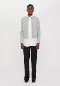 Grå strikket cardigan med knaplukning, prydet med broderidetaljer, over en hvid skjorte med krave og sorte bukser, parret med sorte sko.
