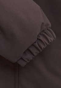 Brunt tyg med en texturerad yta, som har en elastisk manschettdetalj för en tät passform. Materialet verkar hållbart och väderbeständigt.