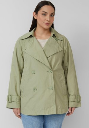 Femme portant un trench-coat vert clair à double boutonnage avec un grand col et des poignets à boucles, associé à un jean bleu, debout devant un fond uni.