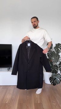 Black coat with notched lapels, single button closure, side pockets, and a smooth interior lining, held by a person in a light shirt.