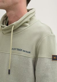 Felpa verde chiaro con cappuccio, presenta un collo con cordino, un pannello superiore strutturato e un pannello inferiore a strisce con il testo blu "#OFF TRACK TOM TAILOR".