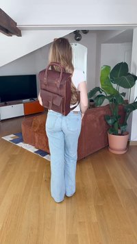 Barna bőrtáska, fedéllel és felső fogantyúval, aranyszínű hardver részletekkel és minimalista dizájnnal, egy személy hátán viselve.