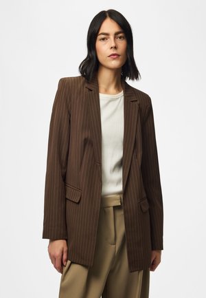 Femme aux cheveux noirs mi-longs portant un blazer marron à fines rayures, un haut blanc et un pantalon beige taille haute, debout devant un fond uni.