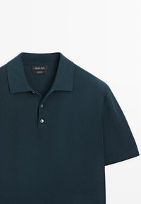 Polo à manches courtes de couleur bleu sarcelle foncé avec col et deux boutons, tissu texturé, présenté sur fond blanc.