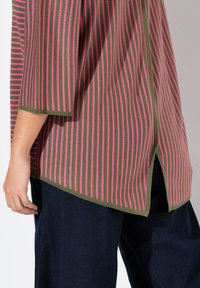 Grünes und pinkes, gestreiftes Strickoberteil mit Dreiviertelärmeln, seitlichem Schlitz und geradem Saum. Kombiniert mit dunkelblauen Hosen.