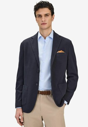 Man klädd i marinblå kavaj, ljusblå skjorta, beige byxor och brunt bälte, med orange näsduk på vit bakgrund.