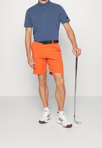 Sininen poolopaita, oranssit shortsit, musta vyö ja valkoiset kengät mustilla yksityiskohdilla. Mies pitelee golfmailaa, seisoo neutraalia taustaa vasten.