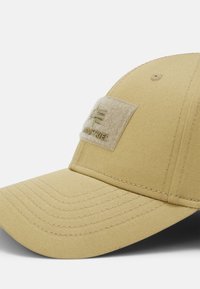 Beige keps med en böjd skärm, texturerad tyg, sömnadsdetaljer och en rektangulär filtpatch på framsidan präglad med en logotyp.