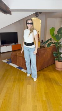 T-shirt bianco aderente, cintura larga nera, jeans svasati azzurri chiari e scarpe bianche; ambiente interno con un divano in velluto a coste marrone e una pianta.