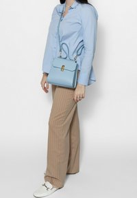  Lichtblauwe leren handtas met een gouden sluiting, hangend aan een strap; gedragen door een persoon in een blauw shirt en beige gestreepte broek.