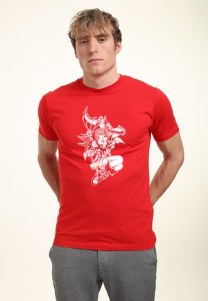 Ung mand med lyst hår iført en rød t-shirt med en hvid tegneseriefigur med hornet hat og støvler på en lys baggrund.