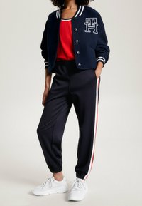 Marinblå bomberjacka med vit randig krage och logotyp, tillsammans med svarta joggers som har röda och vita sidostripes samt vita sneakers.