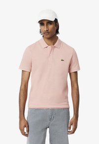 Ljusrosa polo med krage och knapplist med två knappar. Har en grön broderad logotyp. Bärs med ljusgrå jeans och en vit keps.