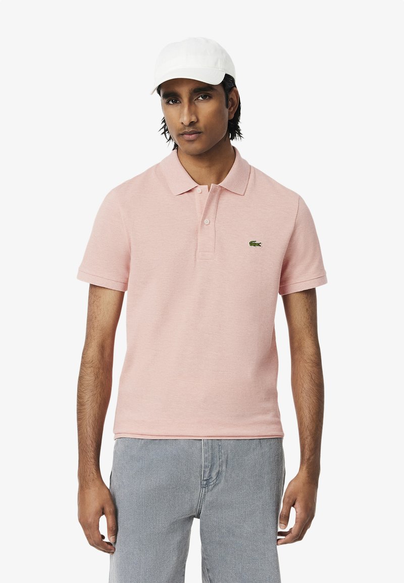 Ljusrosa polo med krage och knapplist med två knappar. Har en grön broderad logotyp. Bärs med ljusgrå jeans och en vit keps.