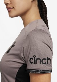 Camisa ajustada de manga corta en tejido gris claro con un patrón texturizado. Presenta detalles en negro y el logo "cinch" en la manga izquierda.