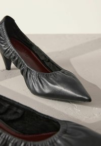 Schwarze Leder-Pump mit hohen Absätzen, spitzer Zehenform und gerafftem elastischen Detail am Schuhkragen. Mit einer tiefroten Innensohle.