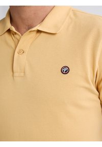 Geel katoenen polo shirt met een gestructureerde weving, een drieknopssluiting en een cirkelvormig logo op de linkerkant van de borst in zwart, wit en rood.