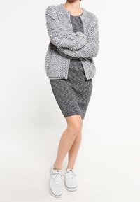 Cardigan en tricot gris flou porté sur une robe gris foncé, associé à des baskets blanches ; mise en valeur de la texture et des éléments de design superposés.