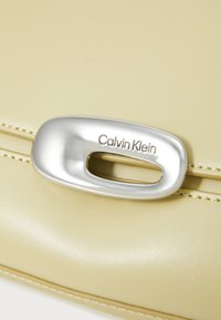 Calvin Klein kott pehmes, heledas kollases nahas; sisaldab hõbedast logo ja tõmblukke ning on nähtava tekstuuri ja õmblustega.