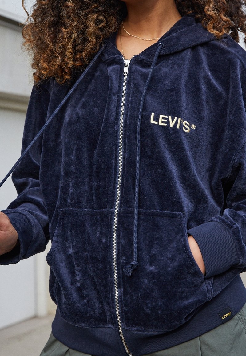 Personne portant un sweat à capuche zippé Levi’s bleu marine avec poches avant et capuche à cordon, debout avec une main dans la poche.