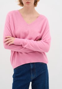 Różowy sweter z dekoltem w serek, z długimi rękawami, ściągaczami na mankietach i u dołu. Połączony z ciemnoniebieskimi dżinsami z klasycznym przeszyciem i kieszeniami.