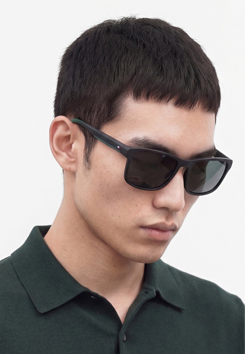 Jeune homme aux cheveux courts noirs portant des lunettes de soleil rectangulaires foncées et une chemise verte foncée à col, regardant légèrement vers le bas.