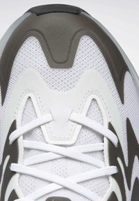 Chaussure de sport blanche avec une tige en mesh, accents gris courbés et lacets blancs. Zone de orteil renforcée et surface à motifs.