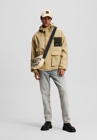 Veste imperméable beige avec une poche en mesh noir, associée à un jean en denim clair et des bottes noires à semelle épaisse. Accessoires comprenant une casquette crème et un sac bandoulière.