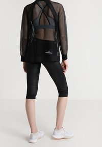 Woman wearing black mesh long-sleeve top, sports bra, black cropped leggings with mesh panels, and white athletic shoes, standing against a plain background.