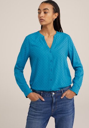 Blusa de botón de color turquesa con puntos texturizados y elevados; mangas largas; escote en V; tejido ligero. Combinada con jeans azules.