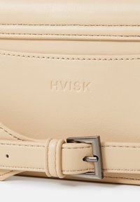 Borsa in pelle beige con logo "HVISK" in rilievo, dettagli cuciti e fibbia metallica sulla tracolla regolabile. Texture liscia.