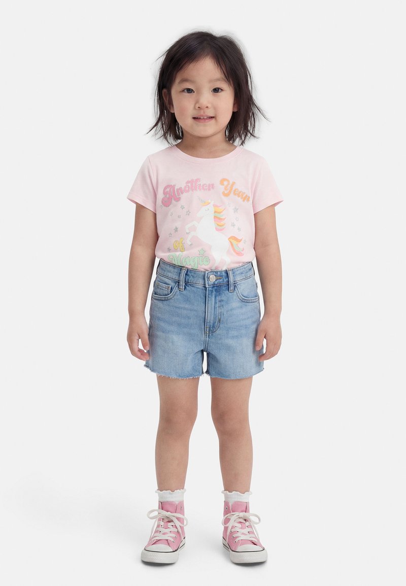 Νεαρή κοπέλα όρθια, φορώντας ροζ μπλούζα με γραφικό μονόκερο, τζιν σορτς, λευκές κάλτσες και ροζ αθλητικά παπούτσια ψηλού τύπου, σε λευκό φόντο.