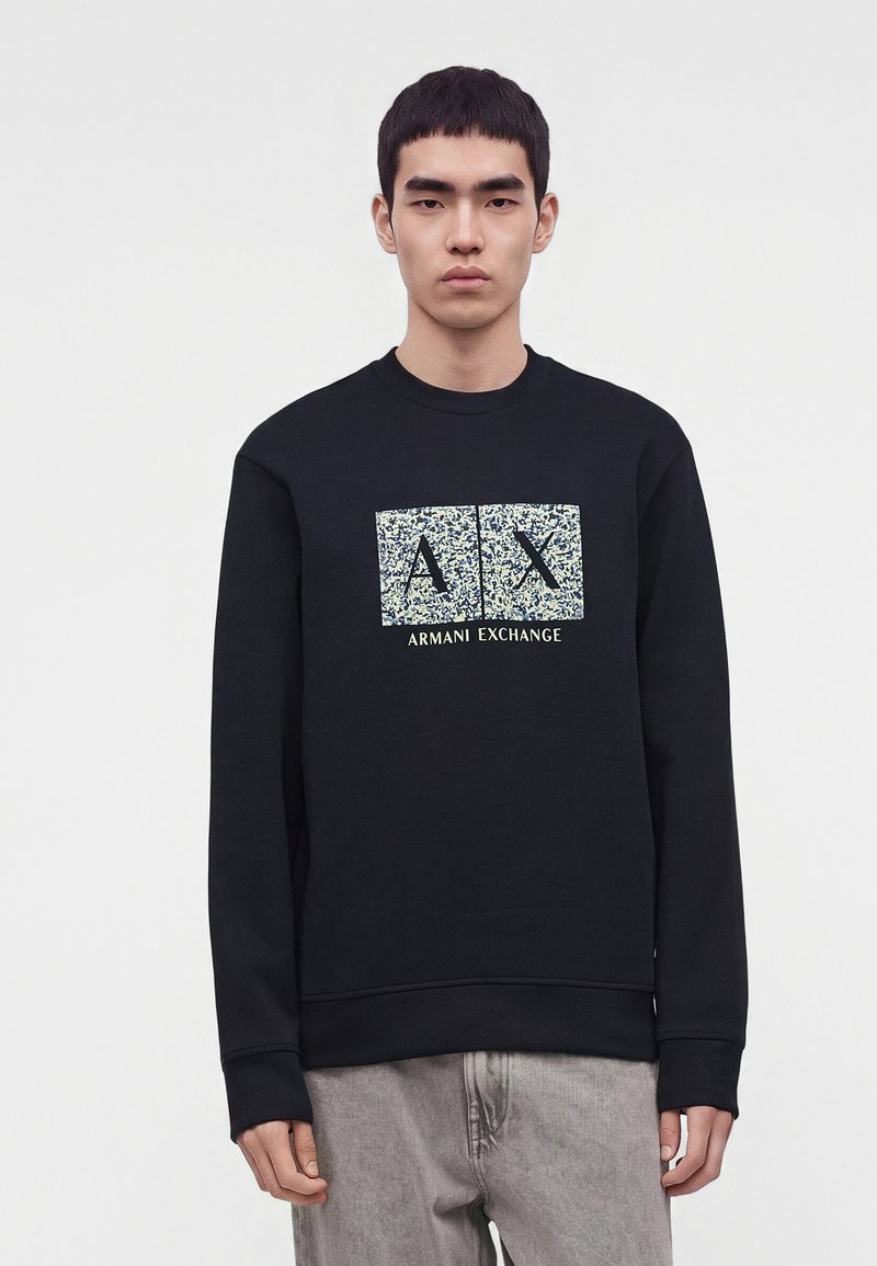 Νέος άνδρας που φοράει μαύρο φούτερ Armani Exchange με υφή λογότυπο AX και ανοιχτό γκρι παντελόνι, στέκεται μπροστά σε απλό λευκό φόντο.