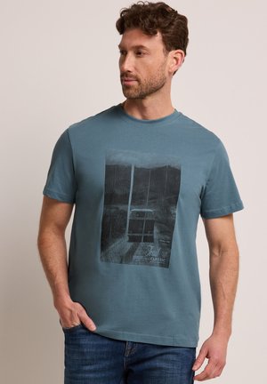 Homme aux cheveux bouclés portant un t-shirt graphique bleu avec une photo en noir et blanc et du texte, debout avec une main dans la poche.