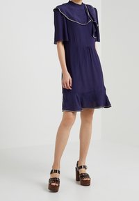 Mörkblå klänning med korta volangärm, hög halsringning, rynkad fåll och kontrasterande detaljer; bärs med bruna platåskor.