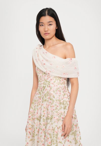 Vrouw met lang zwart haar in een mouwloze, off-shoulder witte jurk met roze en groene bloemenborduursels.