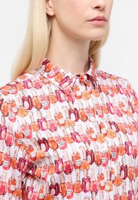 Camicia button-up con un motivo di flaconi di smalto per unghie colorati in tonalità di rosa e arancione su uno sfondo bianco. Materiale in cotone.