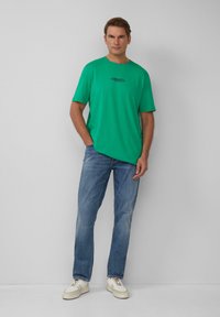 Zielona bawełniana koszulka z krótkim rękawem i okrągłym dekoltem, w połączeniu z wyblakłymi niebieskimi dżinsami i białymi trampkami. Posiada logo na piersi.