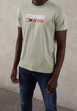 Osoba o ciemnej karnacji ubrana w jasnooliwkową koszulkę Tommy Jeans i ciemnoniebieskie jeansy, stojąca na tle szarej, teksturowanej ściany.