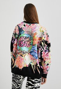 Sweatshirt multicolore avec des impressions florales et architecturales vibrantes ; poignets et ourlet côtelés noirs ; logo "Desigual" bien en vue.