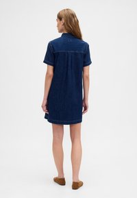 Jeans obleka s kratkimi rokavi, temno modra, z gumbom na ovratniku, ravnim krojem, z všitim robom, nošena z rjavimi semiš copati brez zavezovanja.