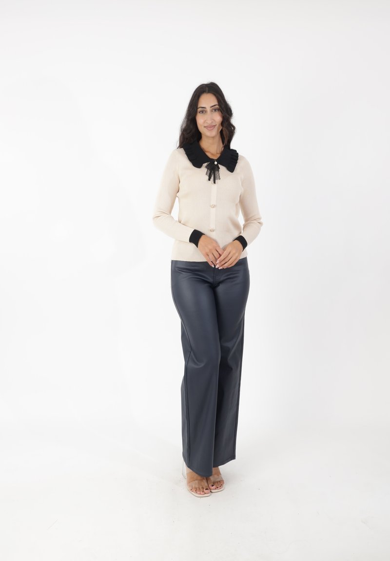 Cárdigan de canalé beige con un cuello negro con volantes y detalle de lazo, combinado con pantalones oscuros ceñidos gris oscuro. El conjunto presenta botones y puños en contraste.