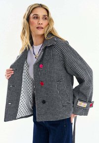 Žena predvádza paleto s čiernobielym vzorom psieho zuba, s farebnými gombíkmi a vnútornou podšívkou so vzorom bielych bodiek, nosí náhrdelník a modré nohavice.