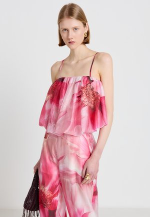 Femme portant un top bustier rose à fleurs et un pantalon large assorti, tenant un sac à main sombre orné de perles, avec des cheveux blonds courts.